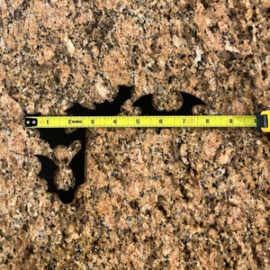 Puede incluir: Dos siluetas de murci&eacute;lagos de metal negro sobre una encimera de granito marr&oacute;n con una cinta m&eacute;trica amarilla que muestra 23 cm.