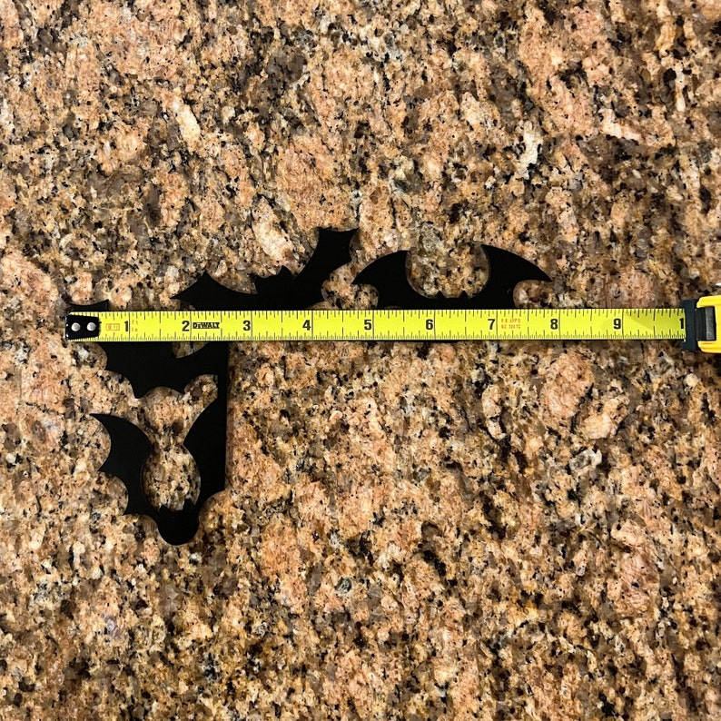 Puede incluir: Dos siluetas de murci&eacute;lagos de metal negro sobre una encimera de granito marr&oacute;n con una cinta m&eacute;trica amarilla que muestra 23 cm.