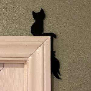 Puede incluir: Silueta de metal negro de un gato sentado en un marco de puerta blanco con la cola extendida por el lado del marco.