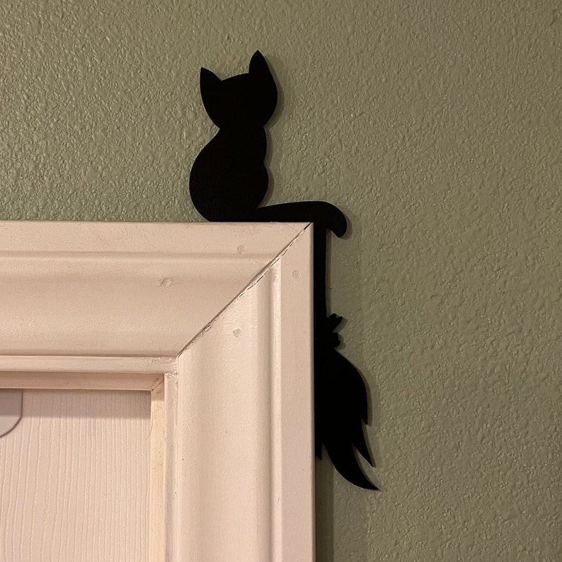 Puede incluir: Silueta de metal negro de un gato sentado en un marco de puerta blanco con la cola extendida por el lado del marco.