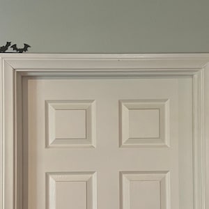 Puede incluir: Una puerta blanca con un dise&ntilde;o de panel elevado, enmarcada por moldura blanca. Se colocan recortes de murci&eacute;lagos negros en la pared sobre la puerta.