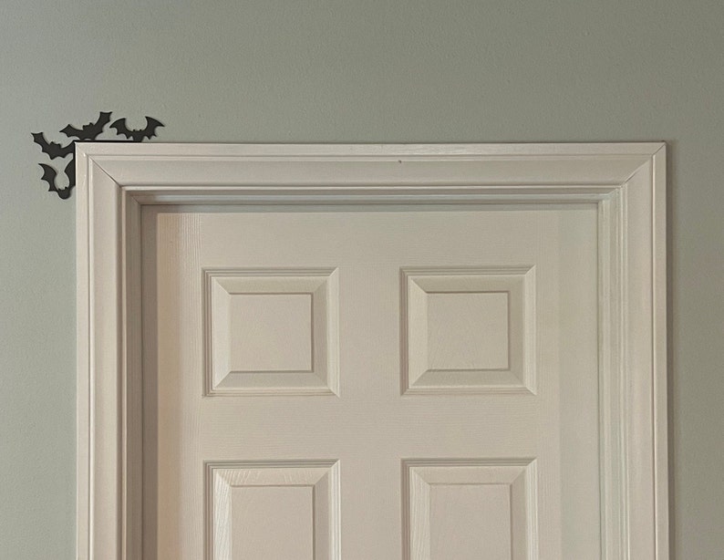 Puede incluir: Una puerta blanca con un dise&ntilde;o de panel elevado, enmarcada por moldura blanca. Se colocan recortes de murci&eacute;lagos negros en la pared sobre la puerta.