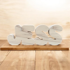 May include: Wooden letters spelling the name "JESS" in a playful, rounded font. The letters are light-colored with visible wood grain, mounted on a small, rectangular wooden base. The background is a light wood surface.