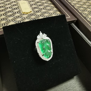 May include: A silver ring featuring a large, vibrant green gemstone carved with a leaf design. The gemstone is surrounded by a halo of small, clear stones. The ring is presented on a black velvet surface, with a decorative box in the background.