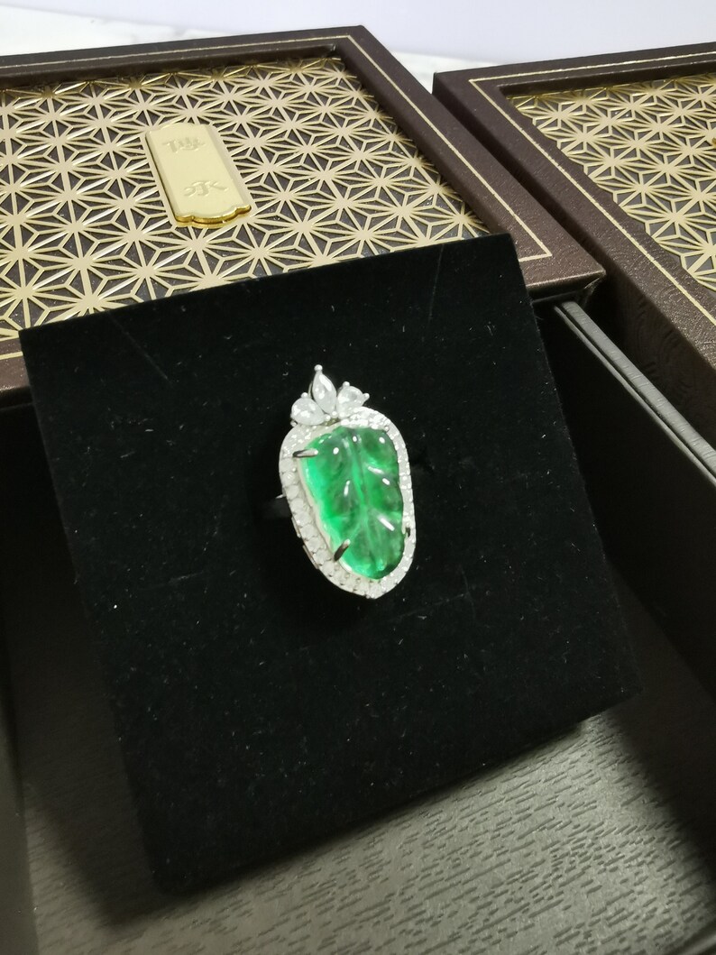 May include: A silver ring featuring a large, vibrant green gemstone carved with a leaf design. The gemstone is surrounded by a halo of small, clear stones. The ring is presented on a black velvet surface, with a decorative box in the background.
