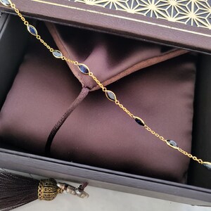 May include: A gold-colored chain bracelet with oval-shaped, dark blue gemstones. The bracelet is displayed inside a dark brown box with a geometric pattern on the lid. A dark brown tassel hangs from the box.