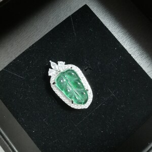 May include: A leaf-shaped pendant with a vibrant green center, framed by a halo of sparkling white stones. The pendant is set on a black velvet surface, suggesting a jewelry display. The design is elegant and detailed.