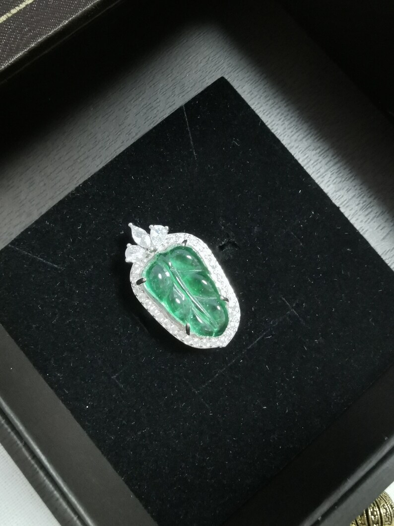 May include: A leaf-shaped pendant with a vibrant green center, framed by a halo of sparkling white stones. The pendant is set on a black velvet surface, suggesting a jewelry display. The design is elegant and detailed.