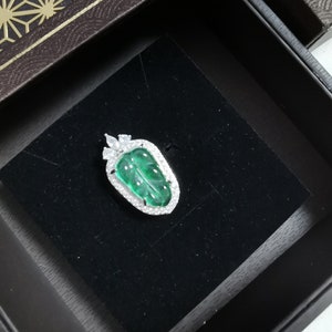 May include: A leaf-shaped pendant with a vibrant green gemstone center, framed by a halo of sparkling white stones. The pendant is set in a silver-colored metal and rests on a black velvet surface inside a decorative box.
