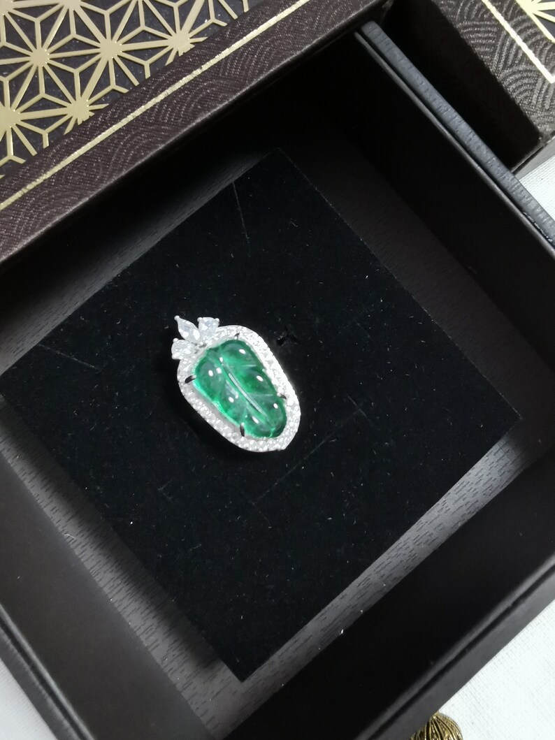 May include: A leaf-shaped pendant with a vibrant green gemstone center, framed by a halo of sparkling white stones. The pendant is set in a silver-colored metal and rests on a black velvet surface inside a decorative box.