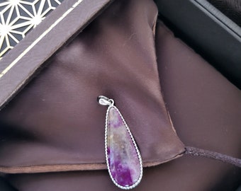 May include: A teardrop-shaped pendant with a purple and tan stone, set in a silver-colored metal frame. The pendant rests on a dark brown satin fabric inside a decorative box.