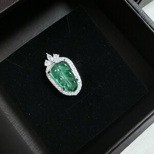 May include: A leaf-shaped pendant with a vibrant green center and a sparkling, clear stone border. The pendant is set in a silver-colored metal and rests on a black velvet surface within a dark box. A unique piece of jewelry.
