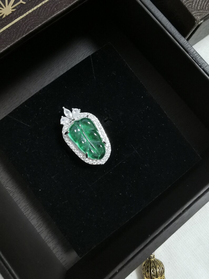 May include: A leaf-shaped pendant with a vibrant green center and a sparkling, clear stone border. The pendant is set in a silver-colored metal and rests on a black velvet surface within a dark box. A unique piece of jewelry.