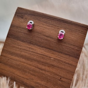 May include: A pair of stud earrings with oval, bright pink gemstones. Each earring is set in silver-coloured metal, with a small decorative accent above the gemstone. The earrings are displayed on a wooden surface.