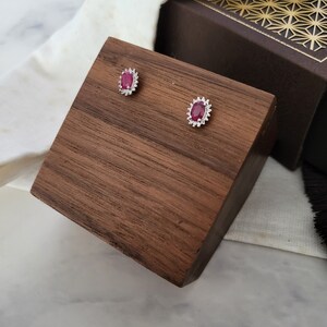 May include: Oval ruby stud earrings with a halo of small, clear stones. The earrings are presented on a dark wooden block. A white cloth and a decorative box are in the background.