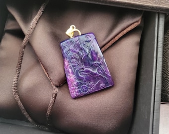Puede incluir: Un colgante rectangular púrpura con un diseño de fénix tallado, suspendido de una fianza dorada. El colgante descansa sobre una tela de satén marrón, dentro de una caja oscura. El fénix está detallado con líneas fluidas.