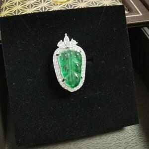 May include: A leaf-shaped pendant with a vibrant green center stone, surrounded by a halo of small, sparkling white gems. The pendant is set on a black velvet surface, showcasing its intricate design and color contrast. The jewelry piece is a statement of elegance.