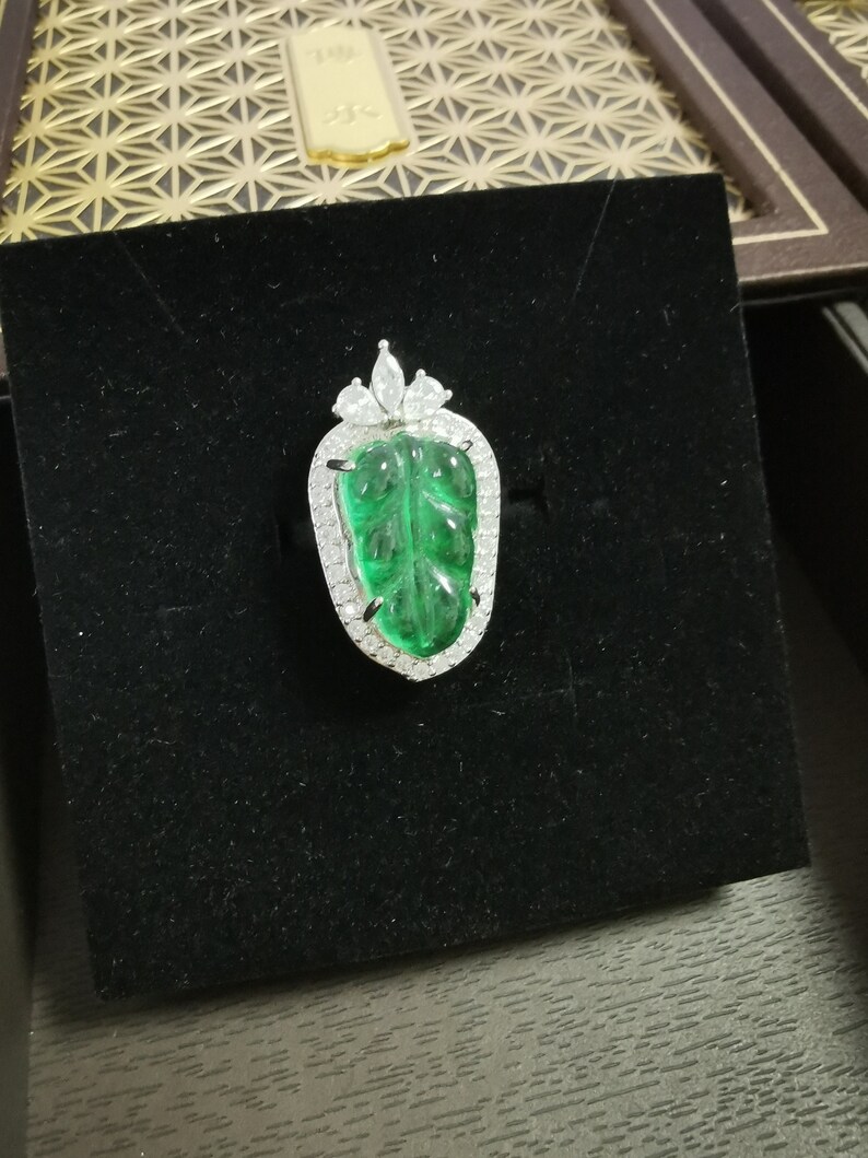May include: A leaf-shaped pendant with a vibrant green center stone, surrounded by a halo of small, sparkling white gems. The pendant is set on a black velvet surface, showcasing its intricate design and color contrast. The jewelry piece is a statement of elegance.