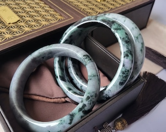 May include: Three jade bangles with a mottled green and grey pattern are displayed in a dark brown box with a gold geometric design. The bangles are smooth and circular, showcasing natural stone variations.
