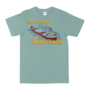 May include: A light blue t-shirt with a graphic of a red and white boat being hit by a large wave. The text "THIS OLD BOAT IS TAKING WATER" is in yellow and orange.
