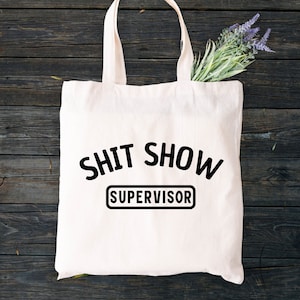 May include: White canvas tote bag with black text that reads "Shit Show Supervisor".