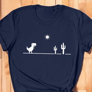 May include: A navy blue t-shirt with a white graphic of the classic dinosaur game. The graphic includes a dinosaur, a sun, and two cacti.