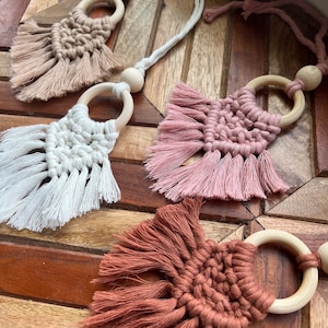 May include: Three handmade macrame wall hangings with wooden rings. The hangings are in shades of brown, pink, and white.