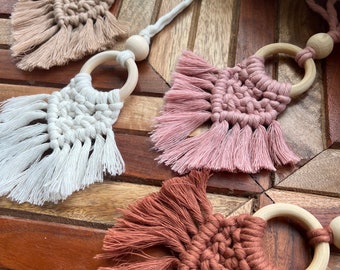 May include: Three handmade macrame wall hangings with wooden rings. The hangings are in shades of brown, pink, and white.