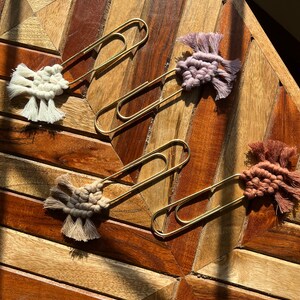 May include: Three gold paper clips with colorful macrame tassels attached. The tassels are in shades of beige, purple, and brown.