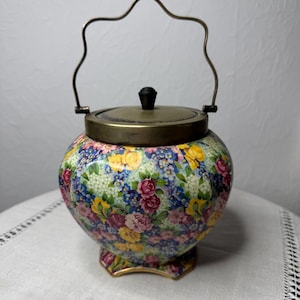 Puede incluir: Una jarra de galletas de cerámica con estampado floral, tapa y asa de latón. La jarra tiene un estampado floral con flores rosas, amarillas y azules sobre fondo blanco.