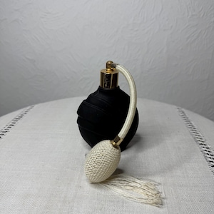 Puede incluir: Botella de perfume de vidrio negro con tapa dorada y atomizador de crochet blanco. La botella está sobre una superficie de lino blanco.