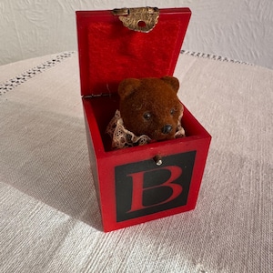 May include: A red wooden music box shaped like a block with the letter "B" on the front. The box is open and a brown teddy bear is inside. The teddy bear is wearing a white lace collar.