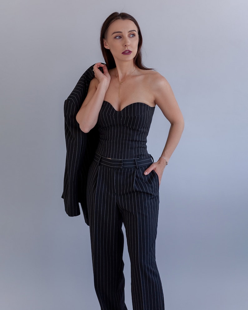 Pode incluir: Uma mulher vestindo um terno de riscas finas preto e branco. O terno inclui um top sem al&ccedil;as e cal&ccedil;as. A mulher est&aacute; segurando um blazer sobre o ombro.
