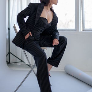 Pode incluir: Uma mulher vestindo um blazer e cal&ccedil;as de terno preto com listras finas senta-se em uma cadeira de prata. Ela est&aacute; olhando para fora da janela.