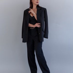 Pode incluir: Uma mulher vestindo um terno preto de riscas finas com um blazer e cal&ccedil;as. O terno tem um caimento cl&aacute;ssico e um estilo moderno.