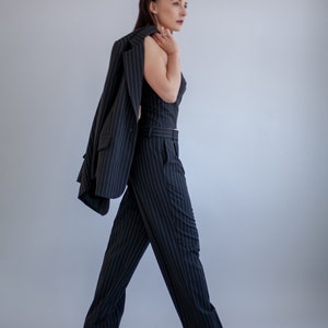 Pode incluir: Uma mulher vestindo um terno de cal&ccedil;as preto e branco listrado com um blazer drapeado sobre o ombro. Ela est&aacute; de p&eacute; com a m&atilde;o esquerda no quadril e a m&atilde;o direita segurando o blazer.