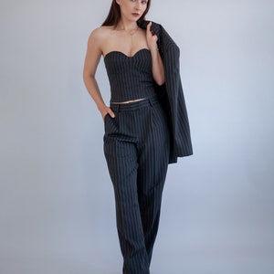 Pode incluir: Uma mulher vestindo um tailleur de cal&ccedil;as de riscas finas preto e branco, com um top sem al&ccedil;as e um blazer drapeado sobre o ombro. Ela est&aacute; em p&eacute; em frente a um fundo cinza claro.