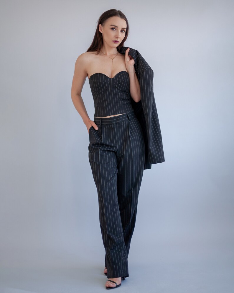 Pode incluir: Uma mulher vestindo um tailleur de cal&ccedil;as de riscas finas preto e branco, com um top sem al&ccedil;as e um blazer drapeado sobre o ombro. Ela est&aacute; em p&eacute; em frente a um fundo cinza claro.