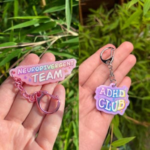 Può includere: Due portachiavi rosa e viola con una catena argentata. Un portachiavi dice "Neurodivergent Team" e l'altro dice "ADHD Club".
