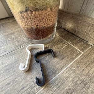 Puede incluir: Dos ganchos en forma de S, uno blanco y otro negro, se muestran sobre una superficie de madera. Los ganchos están hechos de un material sólido y tienen un diseño simple y funcional. Un gran jarrón de vidrio lleno de elementos decorativos está en el fondo.