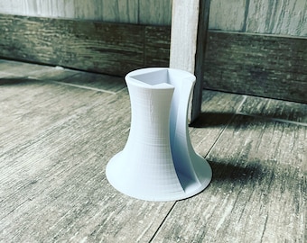 May include: A white, 3D-printed vase with a distinctive, open design. The vase has a flared base and a hexagonal top, with curved sides creating an open space. The vase is on a wooden floor.