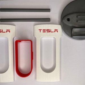 Könnte beinhalten: Eine Sammlung von 3D-gedruckten Tesla-Zubehörteilen. Das Set enthält weiße und rote Komponenten mit dem "TESLA"-Logo, sowie graue zylindrische Stäbe und eine kreisförmige Basis. Die Artikel scheinen für einen bestimmten Zweck bestimmt zu sein.