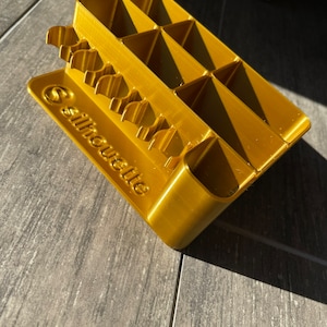 Może przedstawiać: Złoty plastikowy organizer z wieloma przegródkami i logo Silhouette. Organizer jest przeznaczony do przechowywania małych przedmiotów, takich jak materiały do ​​rękodzieła.