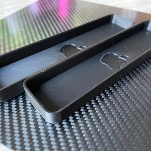 Könnte beinhalten: Zwei schwarze Plastikhüllen, die für Apple TV-Fernbedienungen konzipiert sind. Die Hüllen befinden sich auf einer schwarzen Kohlefaser-Oberfläche.