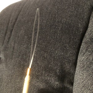 Puede incluir: Una herramienta de color dorado con un alambre delgado y transparente que se extiende desde la parte superior. El alambre está doblado en un bucle en la parte superior.