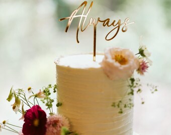 May include: A two-tiered white frosted cake decorated with fresh pink, red, and white flowers. A gold-colored cake topper with the word Always in a cursive script is inserted into the top tier of the cake, standing upright on a thin stem.