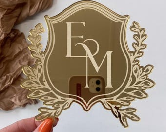 May include: A gold-colored, mirrored acrylic emblem with the letters "E" and "M" in a stylized font. The emblem is shield-shaped, framed by a decorative laurel wreath. The background is a neutral white.