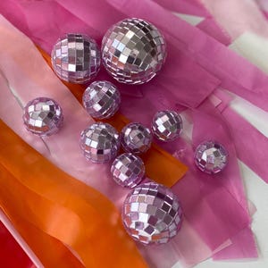 Puede incluir: Una colecci&oacute;n de peque&ntilde;as bolas de discoteca esf&eacute;ricas de color morado claro con facetas de espejo reflectantes, dispuestas sobre un fondo de largas tiras de serpentinas de papel rosa, naranja y blanco, con algunos acentos met&aacute;licos dorados y plateados.