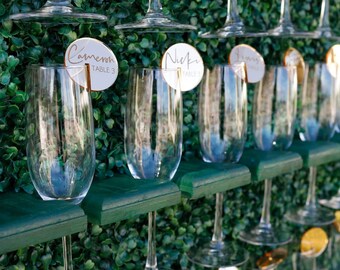 May include: A wall-mounted display featuring rows of clear glass champagne flutes held by dark green wooden shelves. Each glass is adorned with a circular white or gold place card featuring handwritten names and table numbers in black ink.