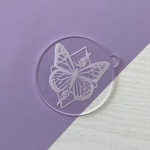May include: A clear acrylic round bauble featuring a white butterfly design. The butterfly has a geometric pattern and is surrounded by leaves.